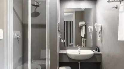 Bagno moderno grigio con doccia, lavabo con specchio, phon a muro e asciugamani bianchi.