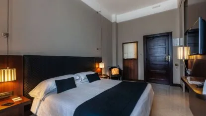 Camera d'albergo moderna con letto matrimoniale, testiera imbottita nera, due lampade sospese e porta in legno scuro.