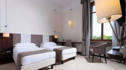 Camera d'albergo moderna con due letti singoli, scrivania, poltrona, ampia finestra e illuminazione soffusa.