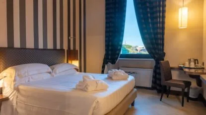 Camera d'albergo luminosa con letto matrimoniale, accappatoi sul letto, poltrona, finestra con tende scure e lampada sospesa.