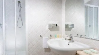 Bagno con piastrelle bianche, lavabo con rubinetto cromato, asciugamano e doccia con parete in vetro.