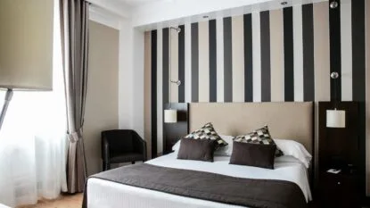 Camera da letto moderna con letto matrimoniale, cuscini decorativi, parete a righe verticali e poltrona nera vicino alla finestra.