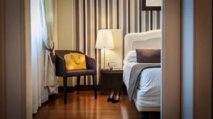 Camera da letto elegante con poltrona nera, borsa gialla, lampada da tavolo, letto con cuscino marrone e scarpe nere sul pavimento.