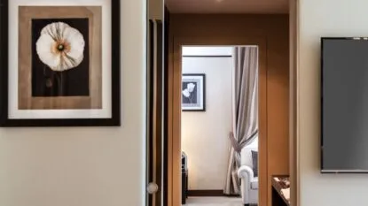 Interno di un soggiorno con porta aperta che mostra un divano bianco, tende grigie e quadri floreali alle pareti.
