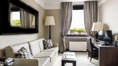 Soggiorno moderno con divano beige, cuscini a quadri, lampada da terra, tavolino, finestra con tende e scrivania con TV.