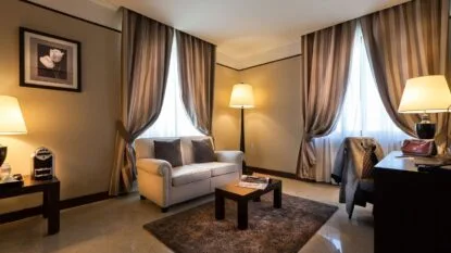 Soggiorno elegante con divano bianco, tavolino, tende a righe, lampade accese e scrivania con sedia e giacca appoggiata.