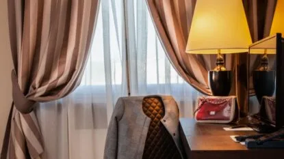 Interno con finestra coperta da tende a righe beige, sedia con giacca grigia e tavolo con lampada, borsa rossa e TV.