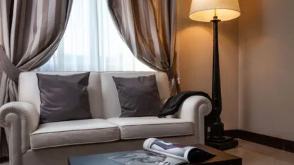 Divano beige con due cuscini scuri, lampada da terra accesa e tavolino con rivista aperta in soggiorno elegante.