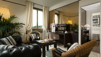 Salotto elegante con poltrone in vimini e pelle, tavolino con vino e snack, piante, lampade e finestra con tende chiare.