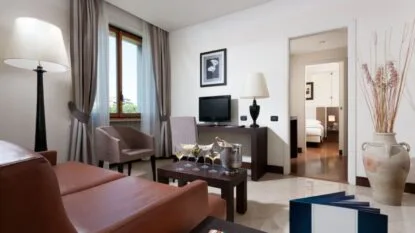 Soggiorno moderno con divano in pelle, poltrona, lampade, tavolino con bicchieri e bottiglia di vino, e vista su camera da letto.