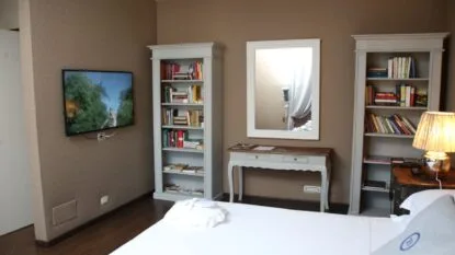 Camera da letto con letto bianco, due librerie grigie, uno specchio e una TV a muro.