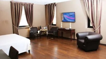 Camera d’hotel spaziosa con pavimento in legno, tre poltrone in pelle marrone, tende violacee e letto con copertina bianca.
