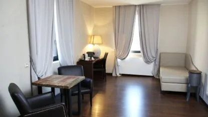Interno di una stanza con pavimento in legno, due poltrone nere intorno a un tavolino, scrivania con lampada e divano beige.