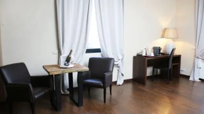 Interno di una stanza con due poltrone nere, tavolo con bottiglia di vino, scrivania, lampada e macchina da caffè.