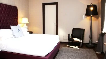 Camera da letto con letto matrimoniale bianco, testiera bordeaux, poltrona nera, lampada da terra e tappeto grigio.