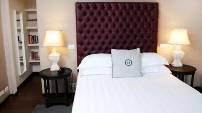 Camera da letto con letto matrimoniale, testiera imbottita bordeaux, cuscino bianco, lampade su comodini e libreria con specchio.