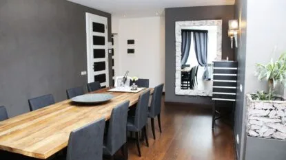 Sala da pranzo moderna con tavolo in legno, otto sedie nere e grande specchio su parete grigia.