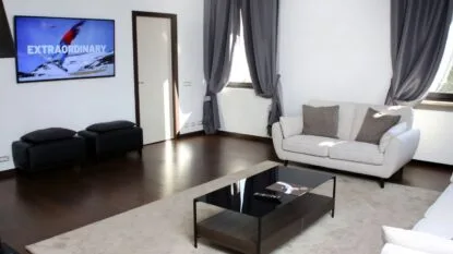 Soggiorno moderno con divani beige, tavolino nero, tende grigie, due pouf neri e televisore a muro acceso.