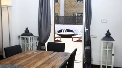 Interno con tavolo in legno e sedie nere, apertura su terrazza con divano tondo e due lettini, lanterne bianche ai lati.