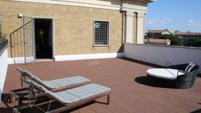 Terrazza con due lettini grigi e un divano rotondo nero con cuscini bianchi, pavimento in legno e muro giallo laterizio.