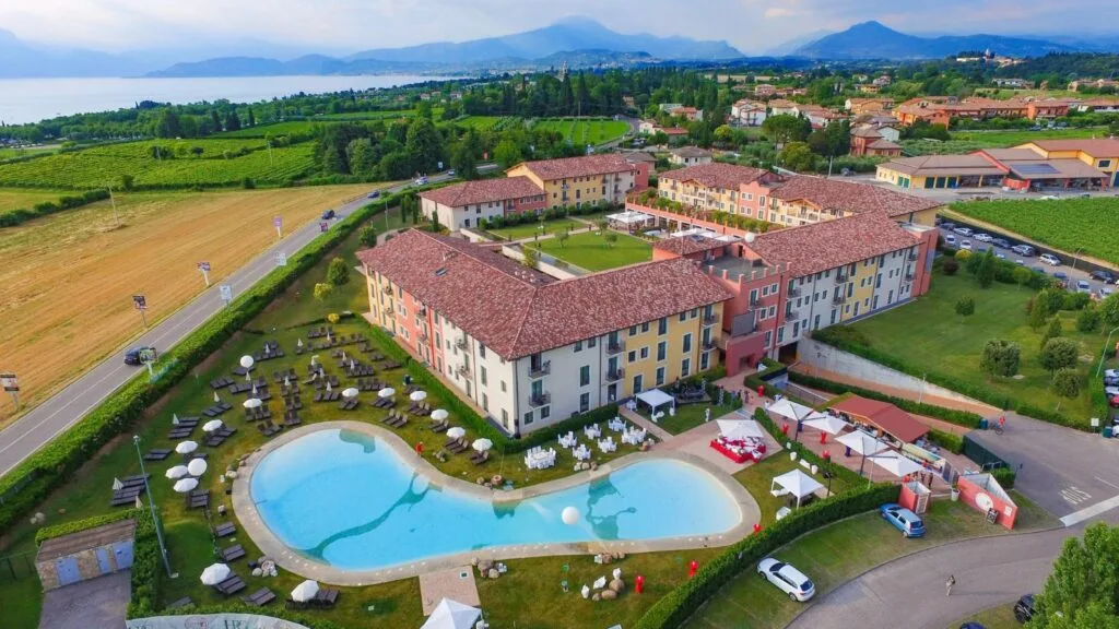 Vista aerea di un complesso residenziale con piscina, tavoli all'aperto e natura circostante, sullo sfondo montagne e lago.