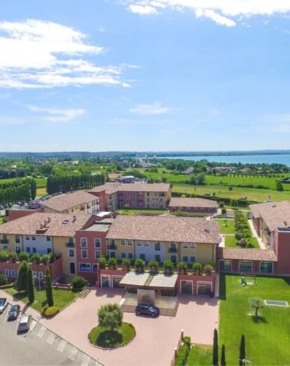 Vista aerea di complessi residenziali circondati da natura e un lago sullo sfondo, sotto un cielo soleggiato.