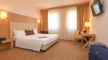 Camera d'albergo moderna con letto matrimoniale, scrivania con lampada, TV a parete e valigie sul pavimento.