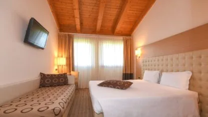 Camera d'hotel luminosa con soffitto in legno, letto matrimoniale, divano con cuscino decorato e TV a parete.