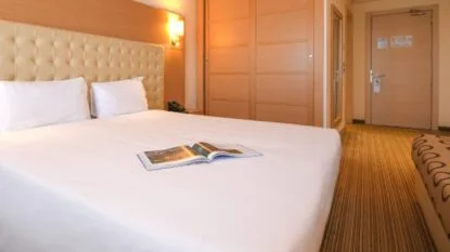 Camera d'hotel con letto matrimoniale, testiera imbottita beige, rivista aperta e armadio in legno chiaro.