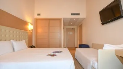 Camera d'hotel con letto matrimoniale, lettino singolo, pareti beige, armadio scorrevole e televisore a parete.