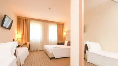Camera d'hotel luminosa con letto matrimoniale, letto singolo, scrivania, televisore a parete e tende beige.