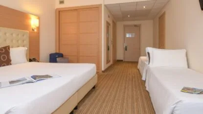 Camera d'albergo con letto matrimoniale, letto singolo, armadio in legno e valigie vicino alla parete.