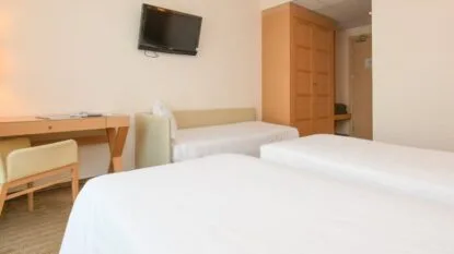 Camera d'albergo con due letti singoli, divano letto beige, scrivania con sedia, armadio e TV a parete.