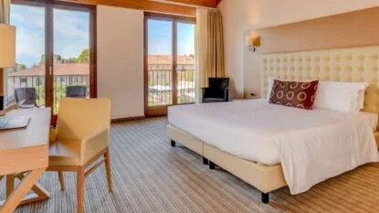 Camera d'albergo luminosa con letto matrimoniale, scrivania, poltrona beige e grandi finestre con vista esterna.