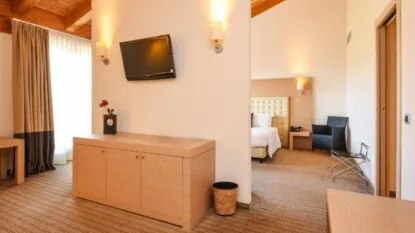 Interno di camera d'hotel con mobili in legno chiaro, letto con testiera imbottita e televisore a parete.