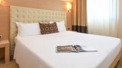 Camera da letto con letto matrimoniale, testiera imbottita, cuscino decorativo e rivista aperta sul letto.