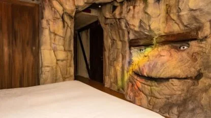 Interno di una stanza con pareti a forma di rocce e un volto dipinto, con letto bianco visibile.