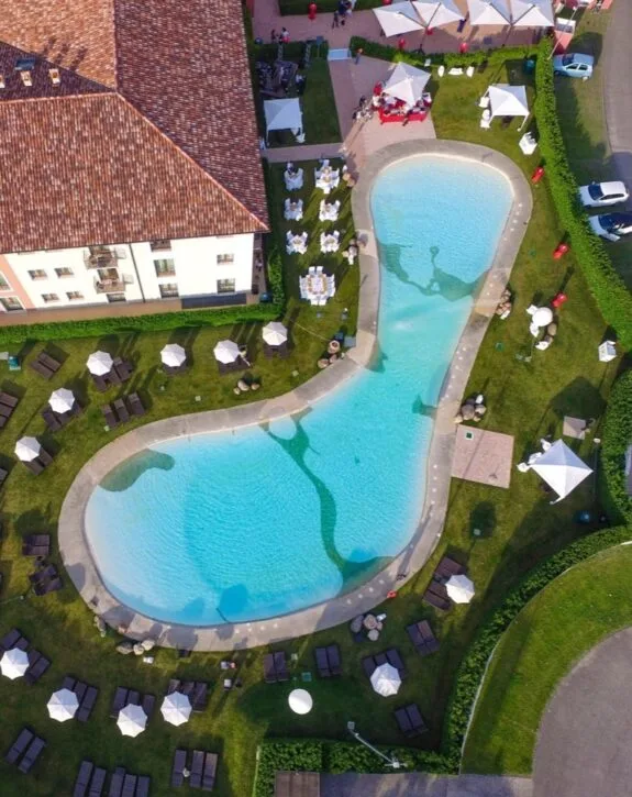 Vista aerea di una piscina a forma di doppio cuore con sdraio, ombrelloni e zone relax intorno in un complesso residenziale.