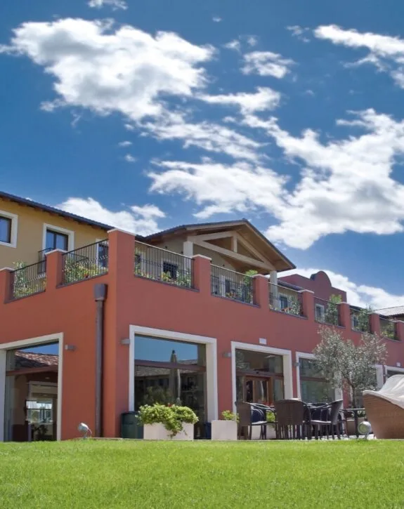 Edificio residenziale con balconi fioriti e zona esterna con tavoli e sedie, sotto un cielo nuvoloso.