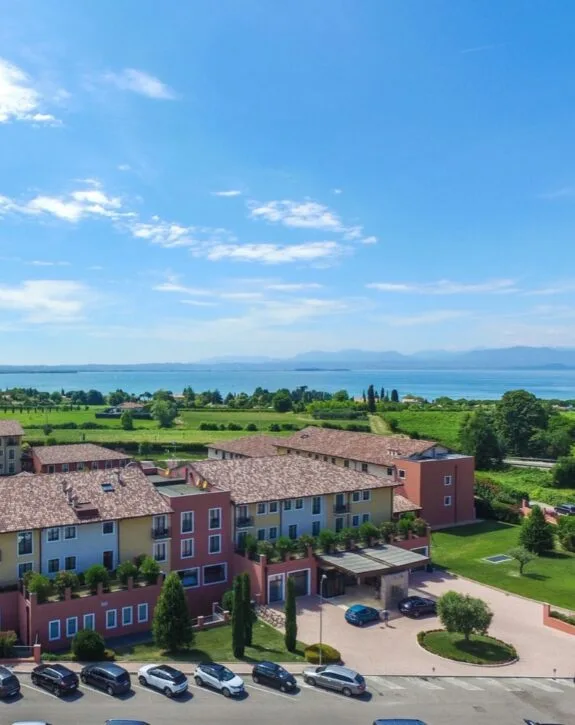 Vista di un complesso residenziale con case colorate, circondato da verde e con un lago e montagne sullo sfondo.