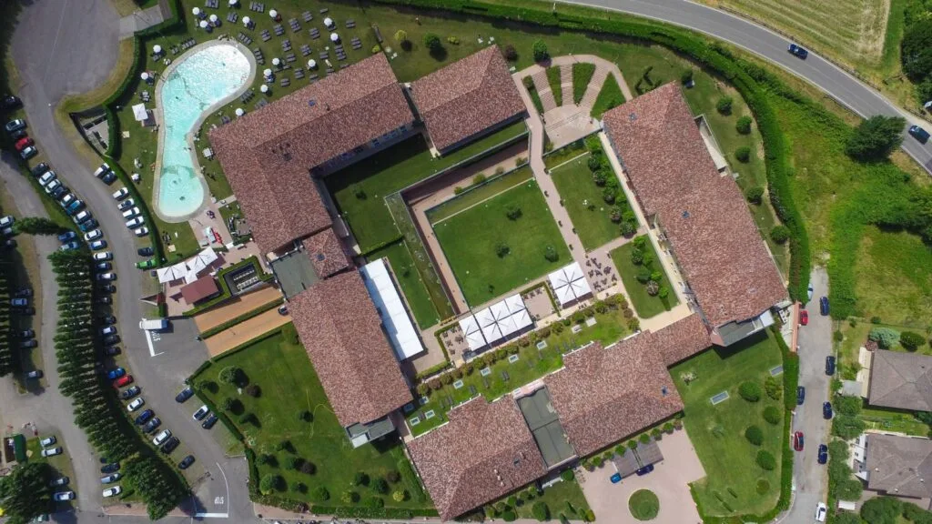 Veduta aerea di un complesso con piscina, giardini e edifici con tetti in terracotta, circondato da strade e parcheggi.
