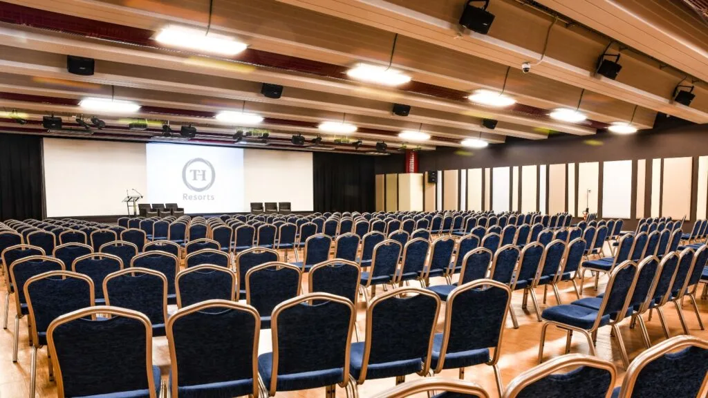 Sala conferenze con sedie blu e un grande schermo con logo "Th Resorts" sullo sfondo.