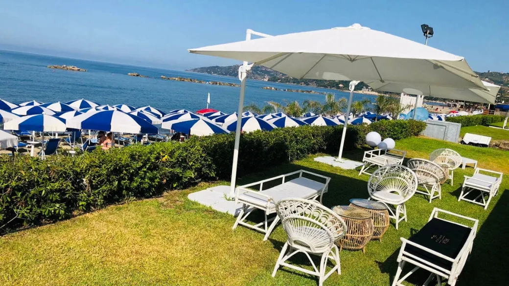 Area lounge sul prato con divani bianchi e ombrellone, vista sul mare e ombrelloni della spiaggia
