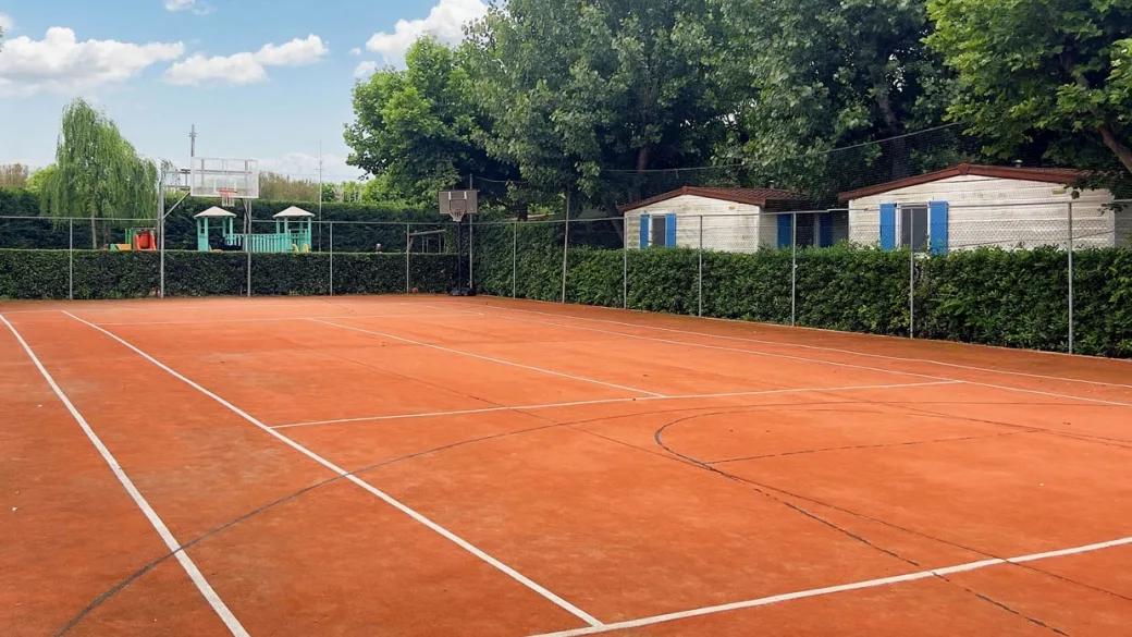 Campo da tennis in terra rossa con area giochi e bungalow sullo sfondo