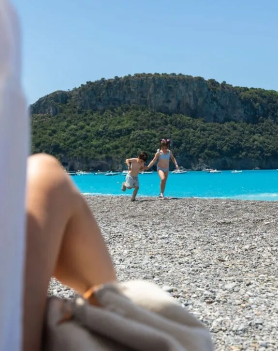 Due bambini si divertono sulla spiaggia con un mare turchese e un grande scoglio verde sullo sfondo.