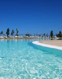 Piscina con acqua cristallina e sdraio verso il cielo sereno con alberi sullo sfondo.
