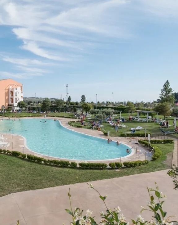 Piscina all'aperto con persone che nuotano, sdraio, alberi e un cielo blu con nuvole.