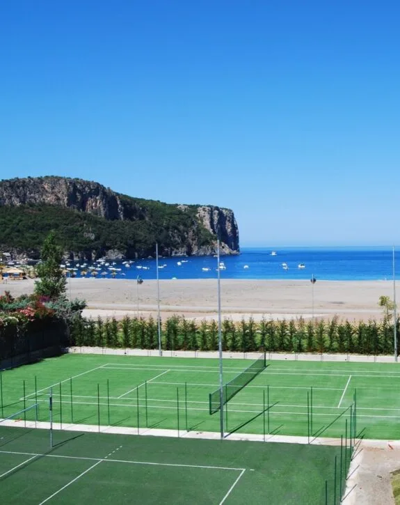 Campi da tennis sul mare con scogliera e spiaggia sullo sfondo, cielo sereno.