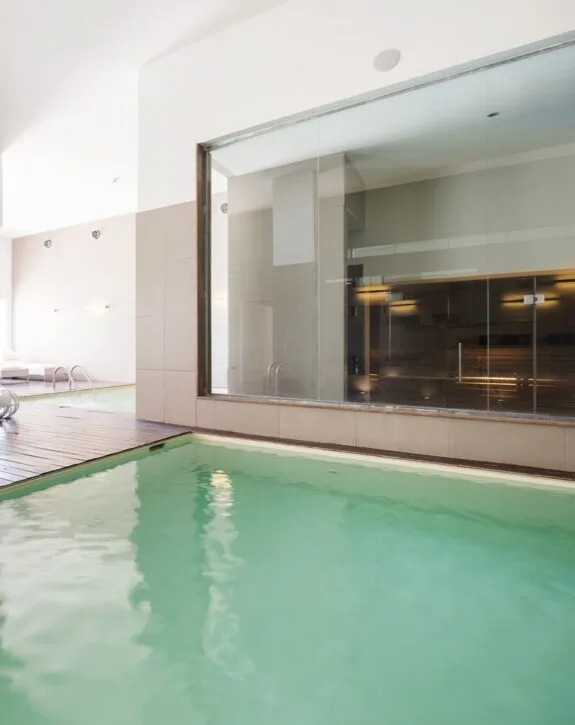 Piscina coperta con pareti di vetro e area relax, ambiente luminoso e moderno.