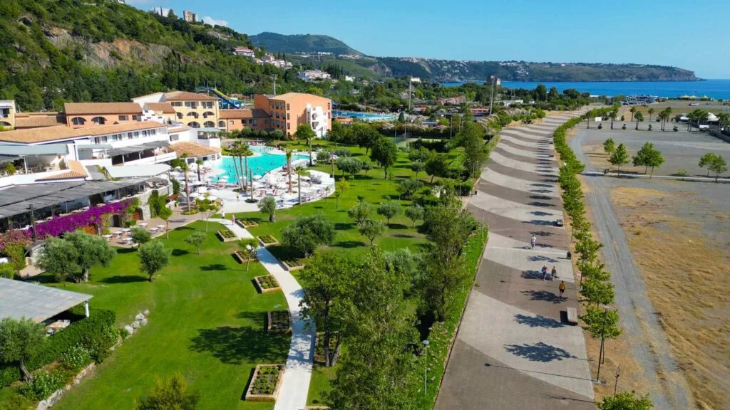 Area pedonale con alberi e panchine vicino a una piscina e strutture alberghiere fronte mare.